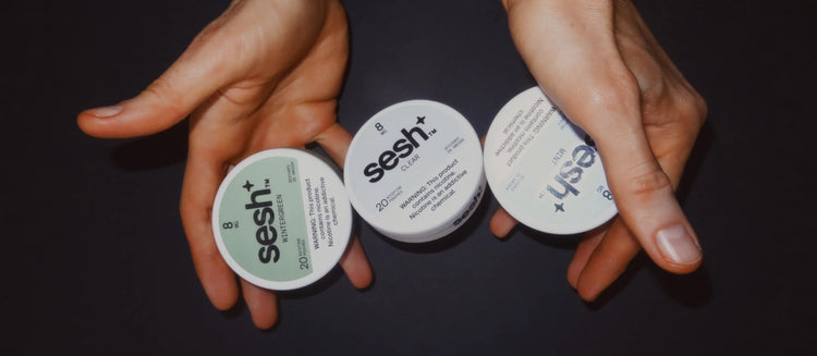 Hands holding three cans of Sesh nicotine pouches in Wintergreen, Clear, and Mint flavors against a dark background.